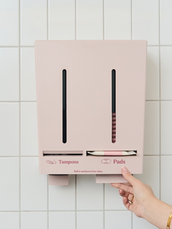 Rosa sanitetsbehållare med fack för tamponger och bindor på en kaklad vägg. En hand når mot dispensern från Herbox.