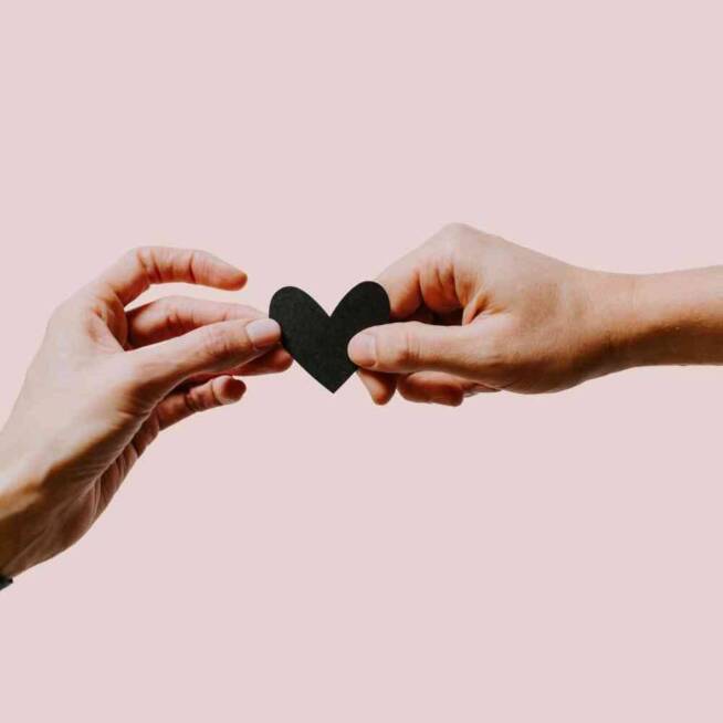 Två händer som håller i ett svart hjärta med rosa bakgrund.