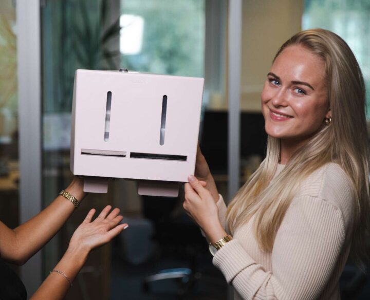 Linn Rosenqvist medgrundare visar upp Herbox mensskyddsautomat i ett kontorslandskap.