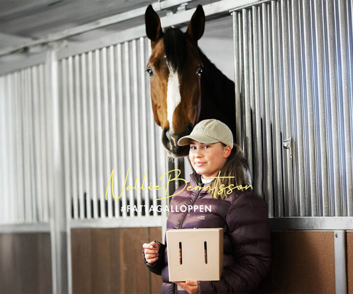 Nellie Berntsson med keps står bredvid en häst i spiltan och håller en Herbox.
