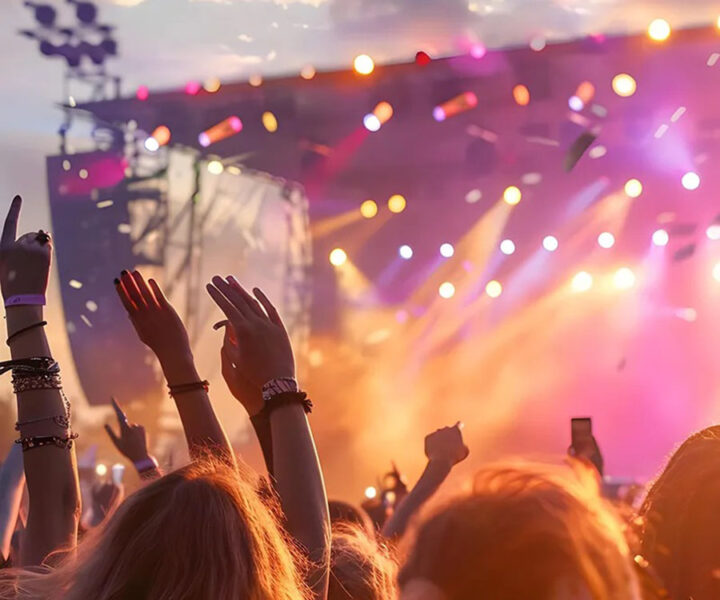 Koncert publikum med hænder i vejret ved koncert med farverige scenelys i baggrunden.