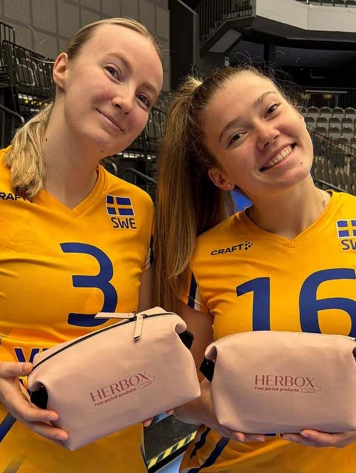 Två leende kvinnor från volleyboll-landslaget i gula tröjor med siffrorna 3 och 16, håller varsin rosa necessär med texten "Herbox".