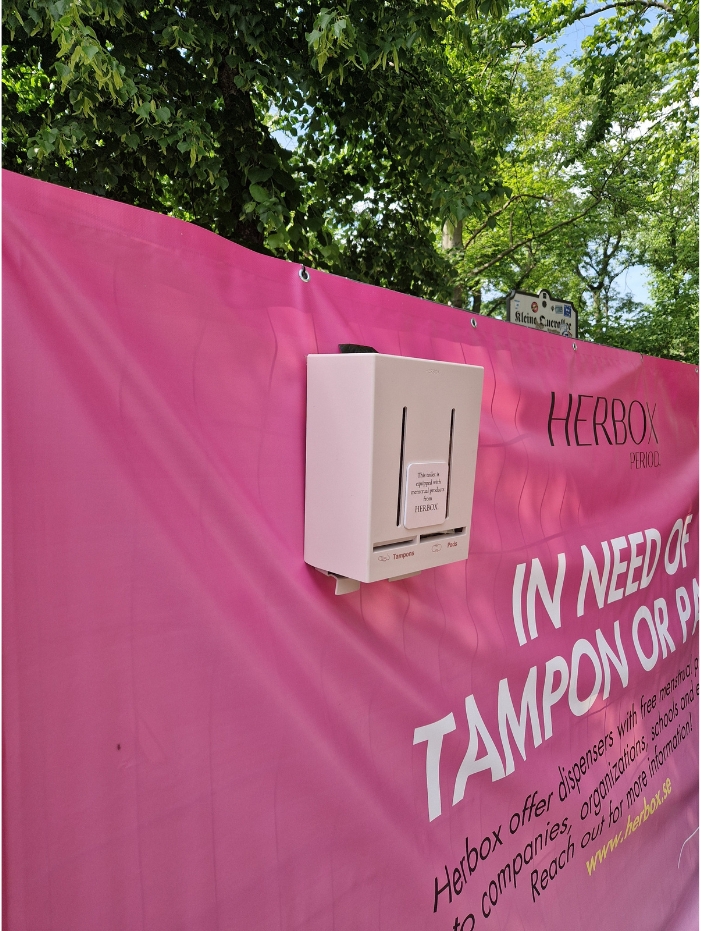En rosa reklambanderoll med en monterad rosa automat för fria mensskydd från Herbox, omgiven av träd.