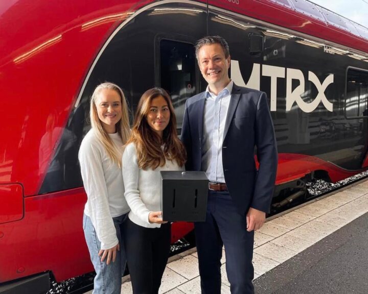 Bild på Linn Rosenqvist och Hanna Lauste grundare av Herbox tillsammans med en representant från MTRX (VR tåg) som visar upp automaterna som finns ombord på alla deras tåg. Med ett rött MTRX tåg i bakgrunden.