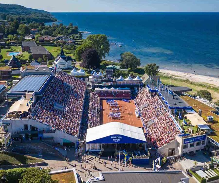 Flygbild av en fullsatt tennisarena vid kusten, med gröna omgivningar och hav i bakgrunden.