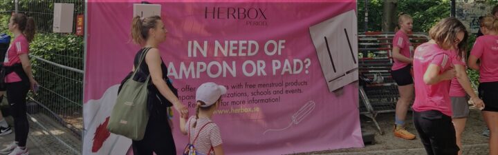 Personer i rosa tröjor framför en rosa banderoll om mensskydd från Herbox.