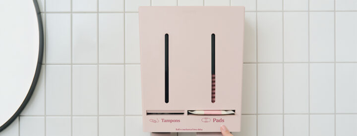 Rosa Herbox låda på kakelvägg, innehåller tamponger och bindor, med en hand som trycker på en knapp.