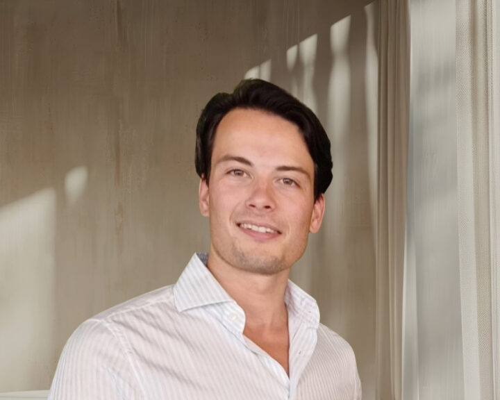 Man in a white shirt smiling in a softly lit room with curtains.