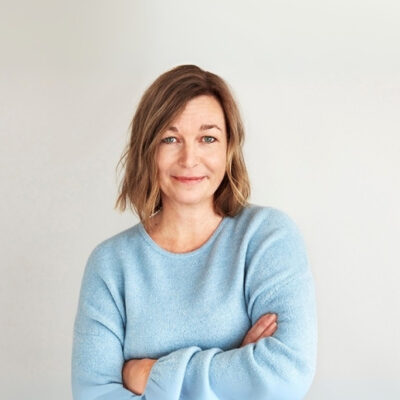 Linda Olsson, Global marknadschef på Lindex med brunt hår i blå tröja, armarna i kors. Ljus bakgrund.