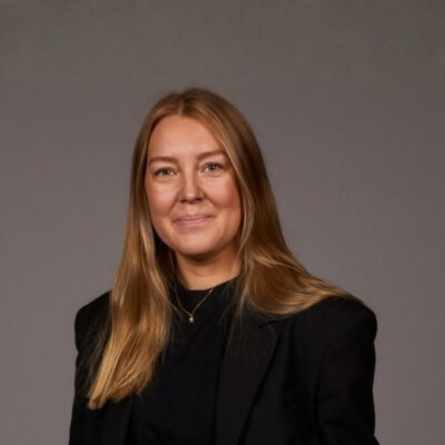 Linnéa Nilsson Junior HR-partner, Göteborgs symfoniker med långt hår i svart kavaj mot grå bakgrund.