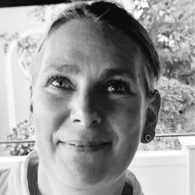 Mia Jonasson Avdelningschef medarbetarservice på Liseberg i svartvitt, ler med bakgrund av träd och byggnad.