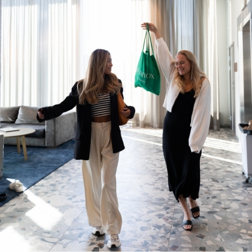 Twee vrouwen binnen, lachend en wandelend, één vrouw houdt een groene tas omhoog.