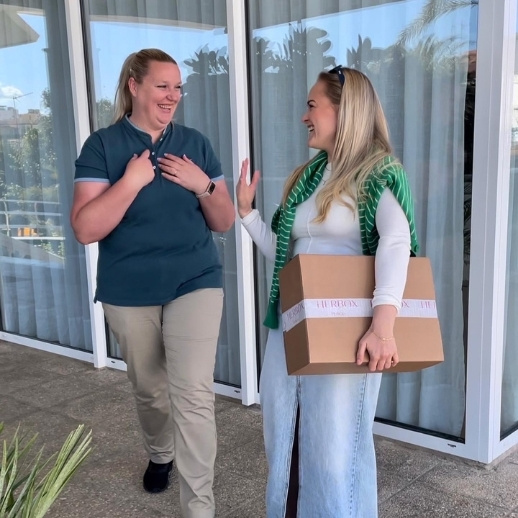Twee vrouwen lachen en praten buiten, terwijl een van hen een doos vasthoudt.