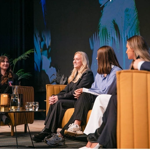 Groep vrouwen in gesprek op een podium met comfortabele stoelen, glazen water en een kunstzinnige achtergrond.