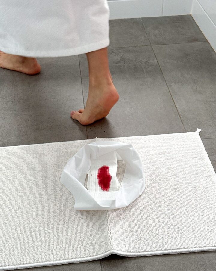A person walking on a grey tile floor, with a white towel, sanitary pad with red stain on a white mat.