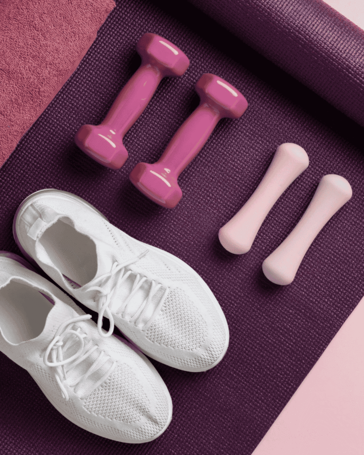 White trainers, pink dumbbells, and a purple yoga mat arranged on a pink and purple background.