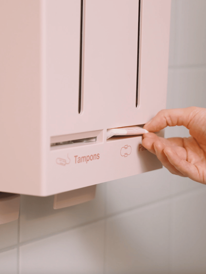En hand tar en tampong från en rosa automat med texten "Tampons" och "Pads" på en vägg i ett badrum.