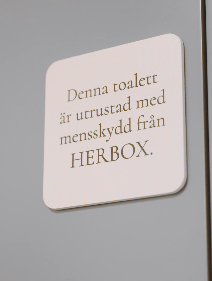 Skylt på en toalett som informerar om att mensskydd från HERBOX finns tillgängligt.