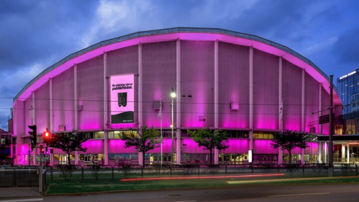 Stort arenahus upplyst i rosa ljus på kvällen, träd och gata i förgrunden, molnig himmel.